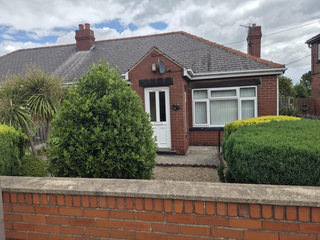 A red brick bungalow with large front window. The gravelled front garden includes several shrubs and bushes and two palm trees.