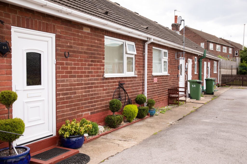 A red brick bungalow within a small terrace of bungalows. Small bushes and plants in pots line the outside of the bungalow.